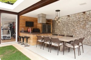 Anexo para área gourmet com estrutura em aço corten e parede de pedra moledo. Madeira de demolição nos painéis e nos armários sob o tampo de porcelanato Concrete Brown.