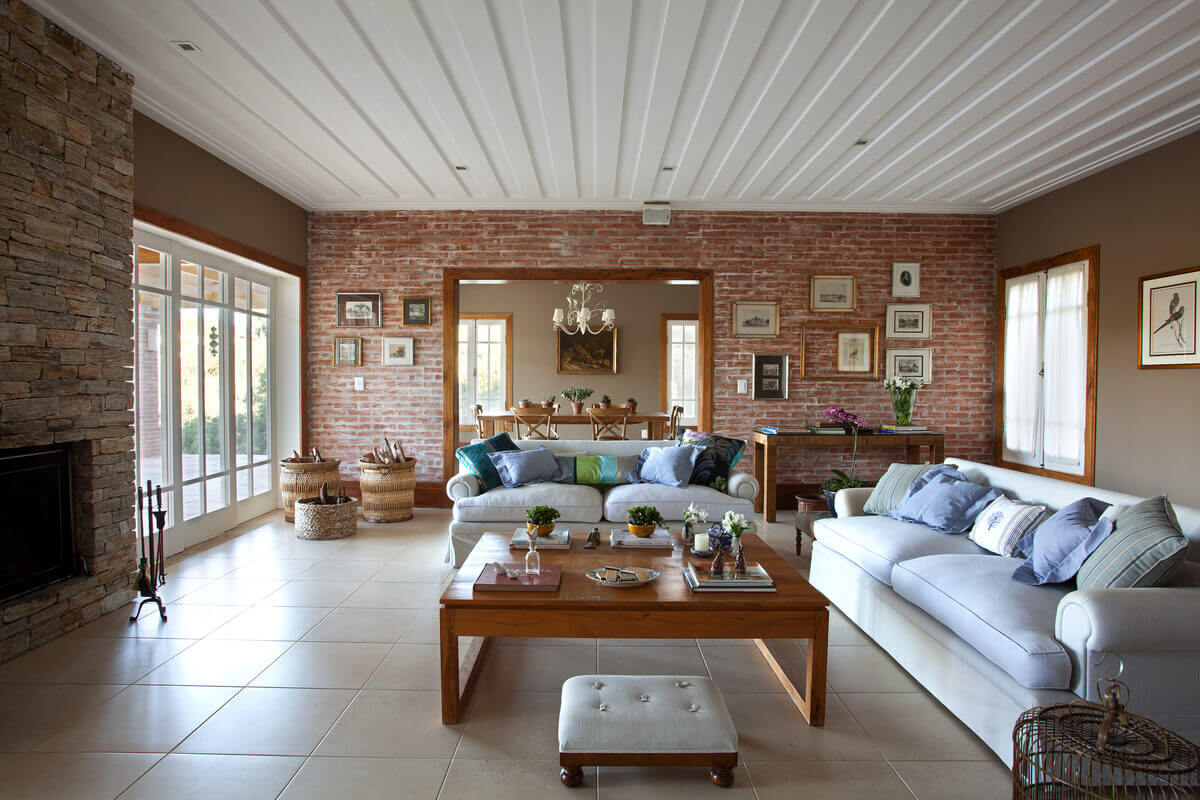 Sala de estar com paredes de tijolos rústicos, lareira de pedra madeira e forro de gesso saia e blusa.