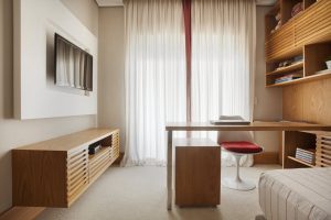 Quarto masculino com escrivaninha em freijó e painel de TV bege com toques de vermelho.