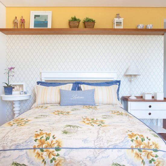 Colcha e porta travesseiros em tecidos da Entreposto. Cabeceira revestida com papéis de parede separados por prateleira decorativa.